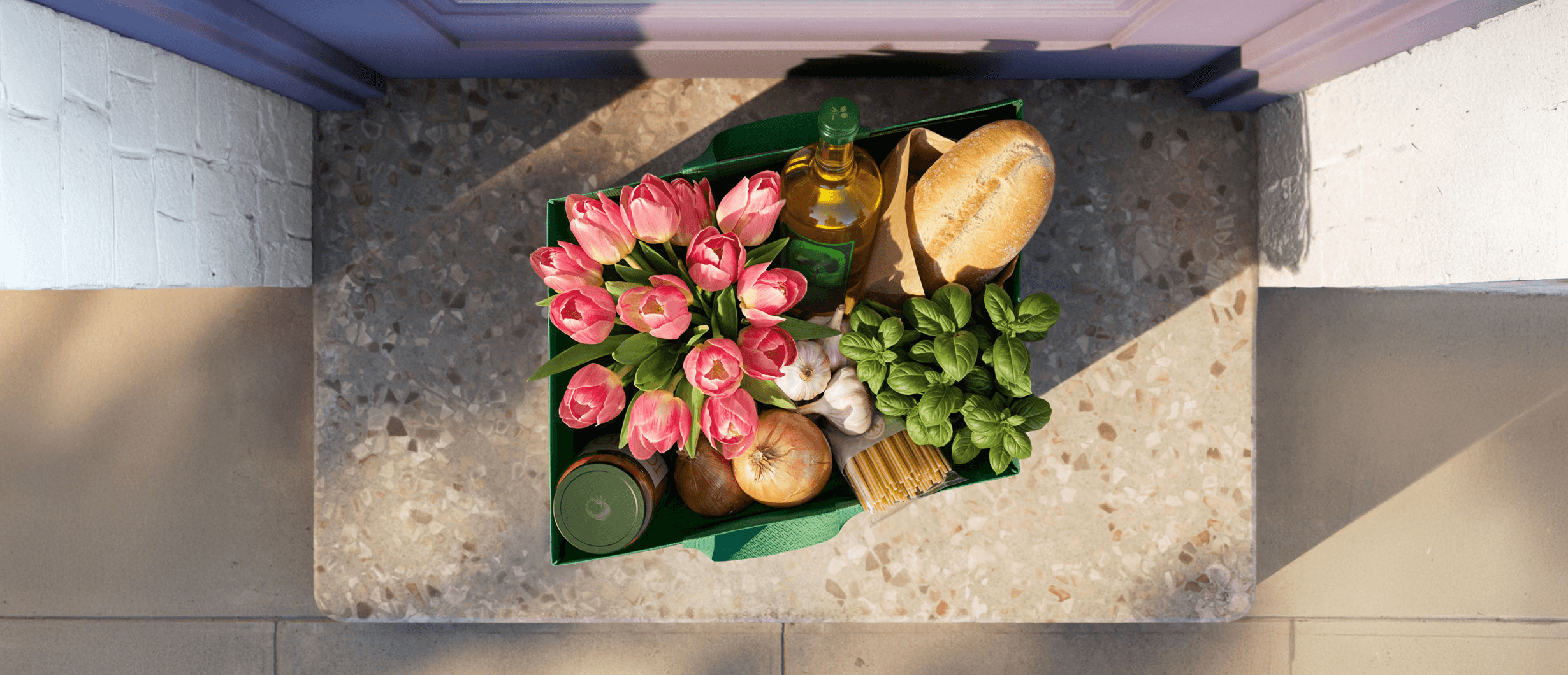 Overhead view of a door stoop with a grocery delivery bag containing flowers, bread, olive oil, and produce, where the bag's shape forms Stripe's parallelogram logo.